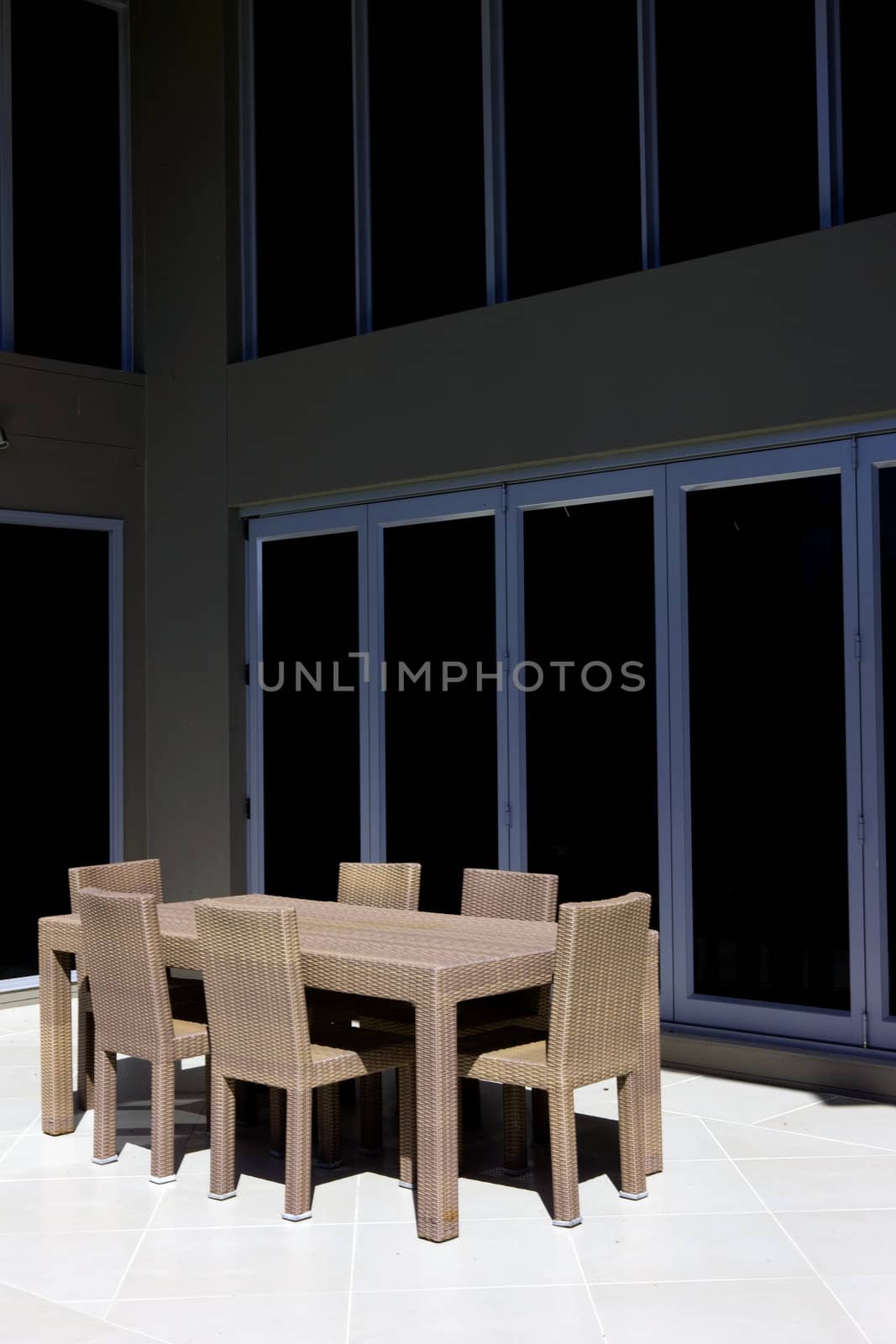 Empty table and chairs standing in th sunshine on the porch of an upmarket residence in front of large glass doors or windows