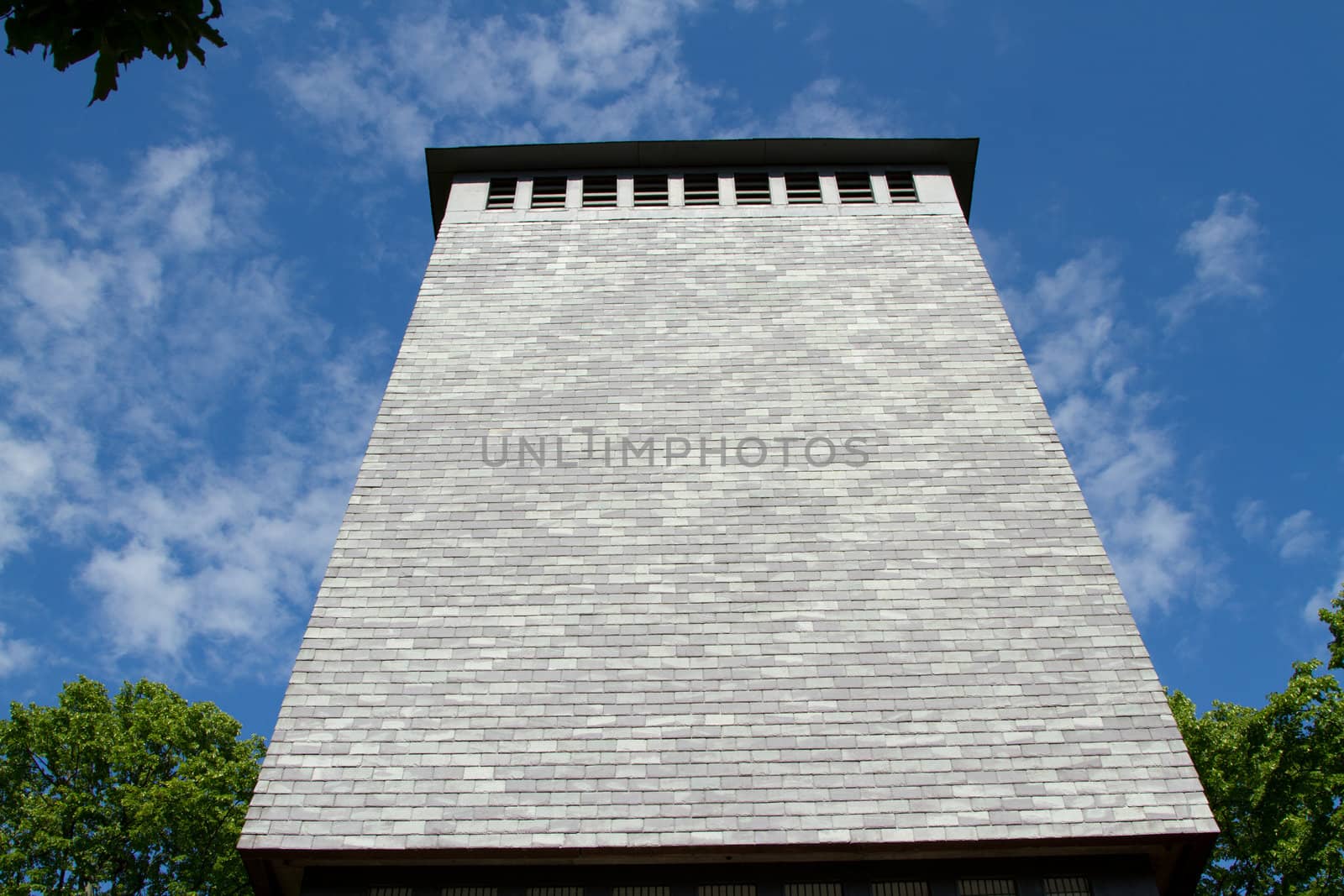 A tower facade covered in slate tiles rises to windows and a roof with green trees and a blue sky.