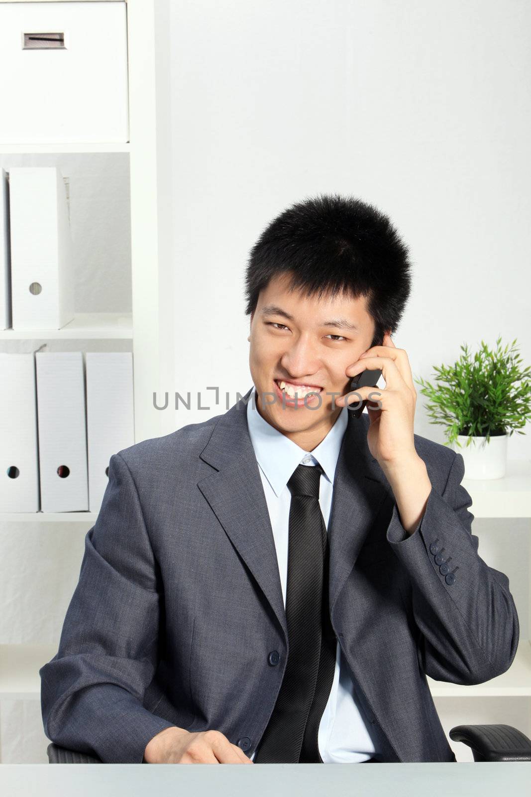 Young Asian businessman seated at his desk talking a cellphone