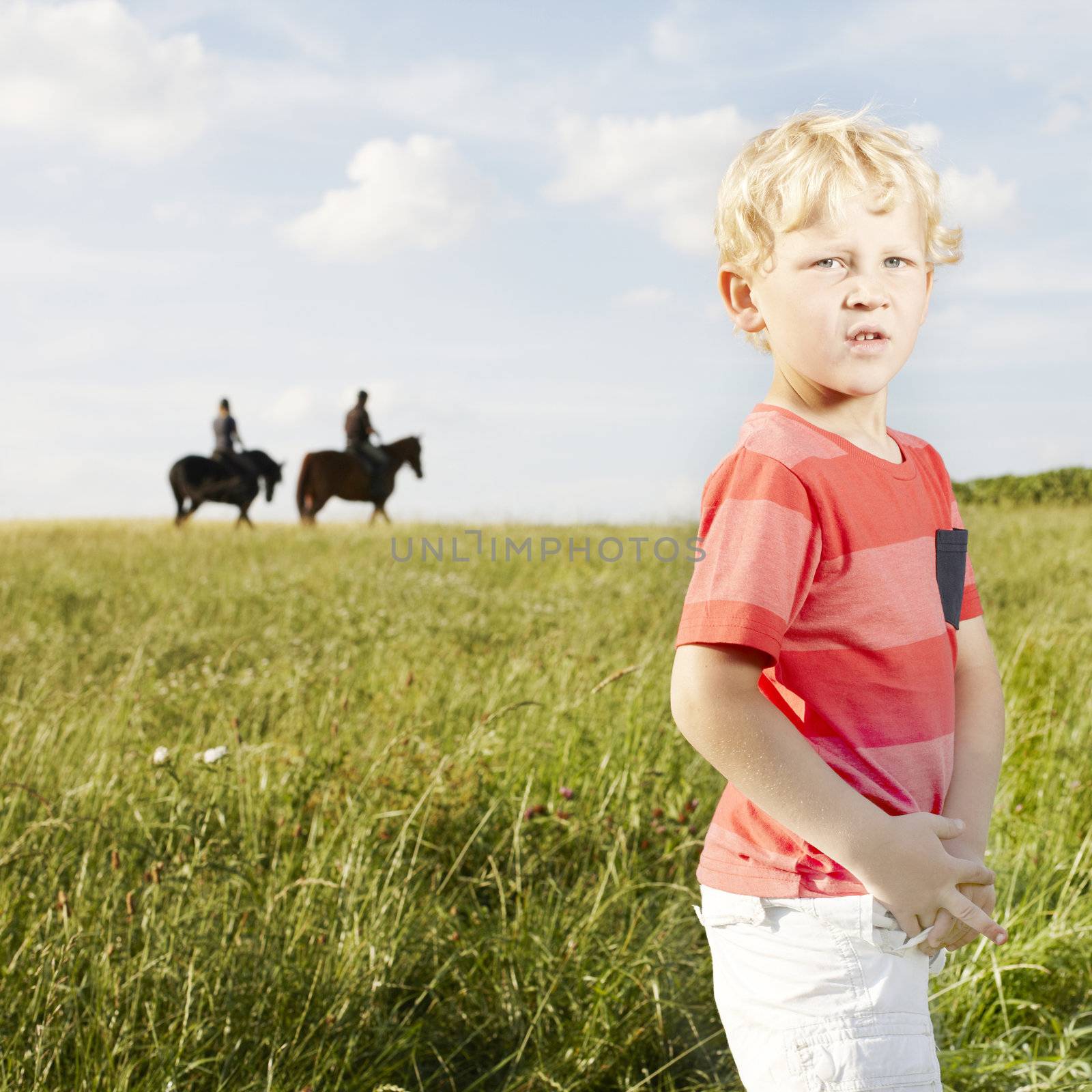 Young blonde boy standing in a grassy field with two horse riders silhouetted on the skyline in the background