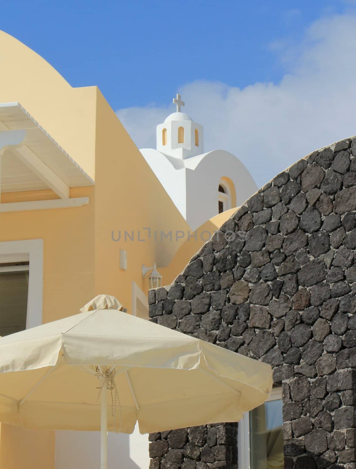 Small white dome of an orthodox church behind orane house and black wall with umbrella at Santorini, Greece