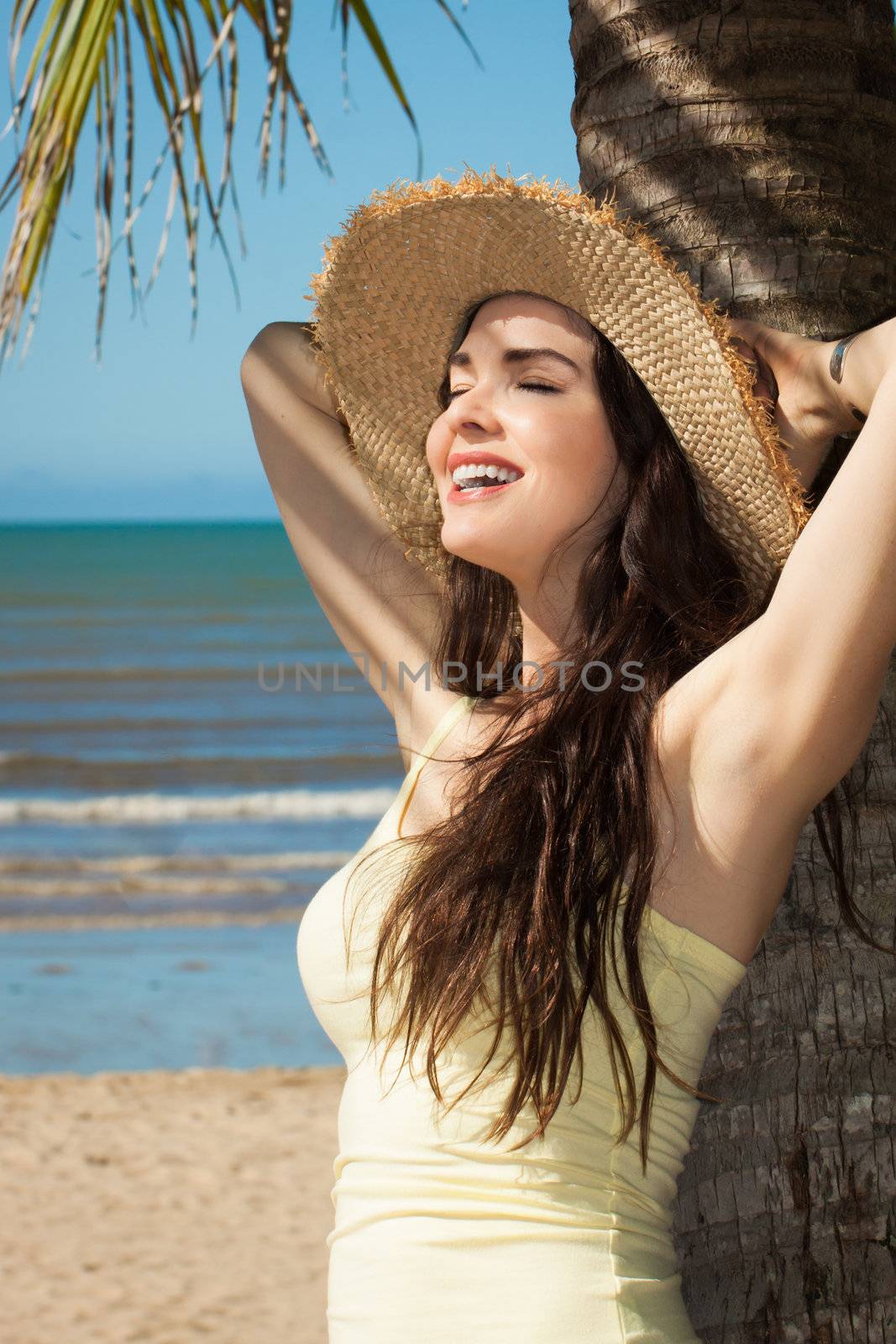 A beautiful young woman leaning against palm tree on a tropical beach wearing a straw hat.