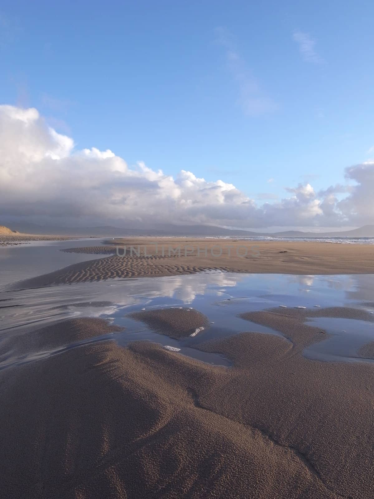 A sandy beach with rippled sand and a dune with the tide bringing the sea over the sand with reflections of a blue cloudy sky.