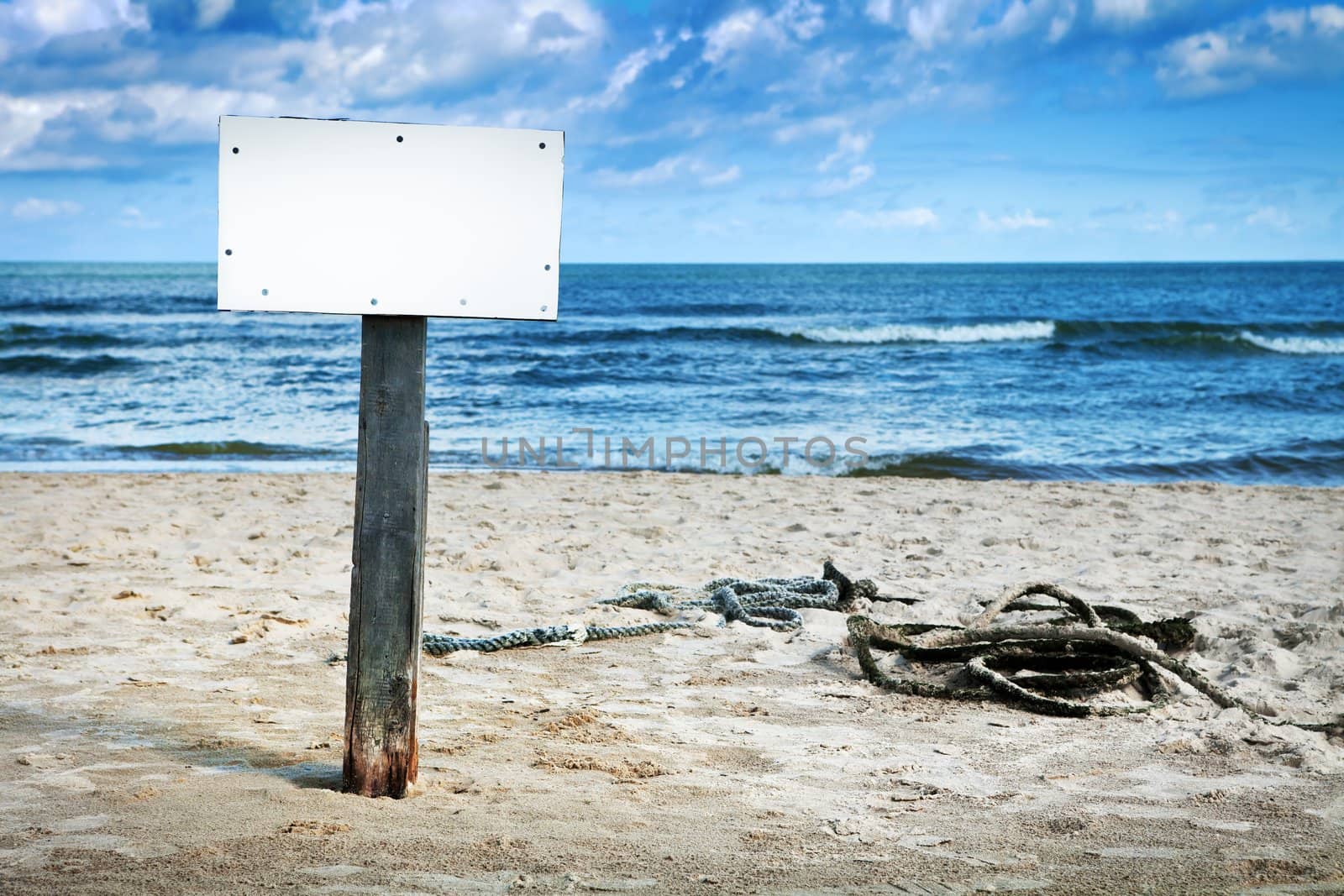 Blank white board on wooden pole on sandy beach, cloudy sky and sea in background