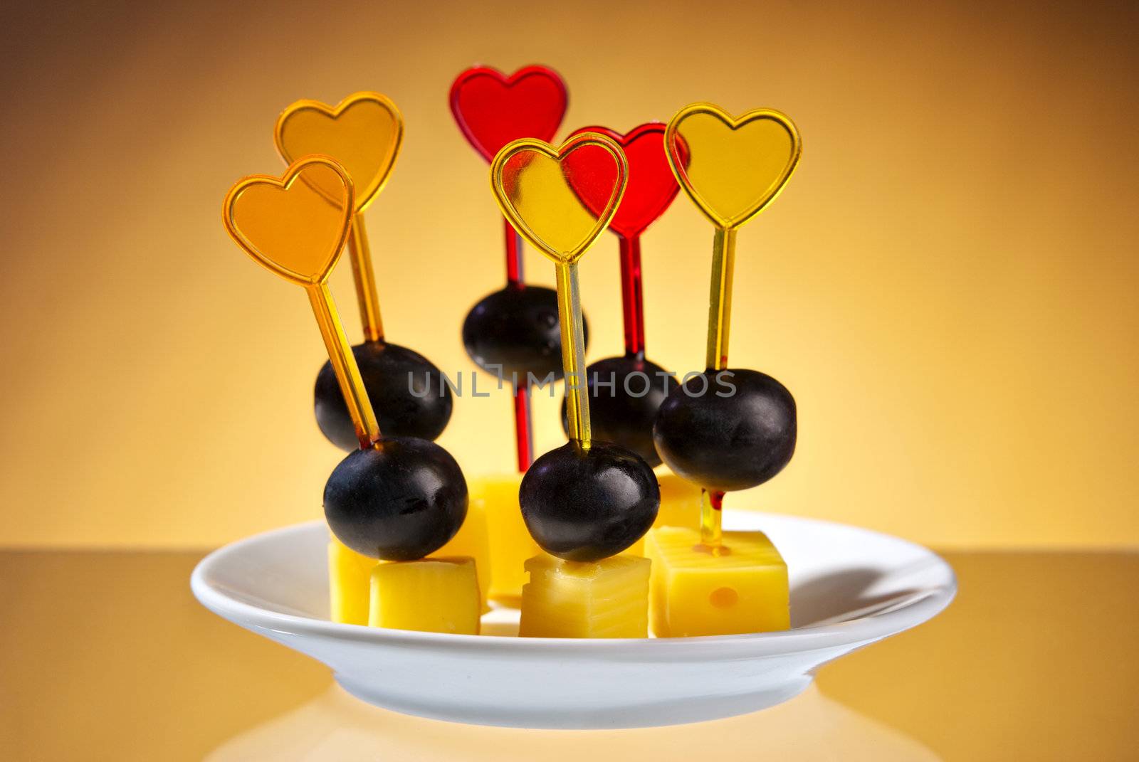 Closeup of delicious cheese and grape canape sandwich made from cheese, black grapes and grape threaded on heart shaped toothpick over yellow gradient  background