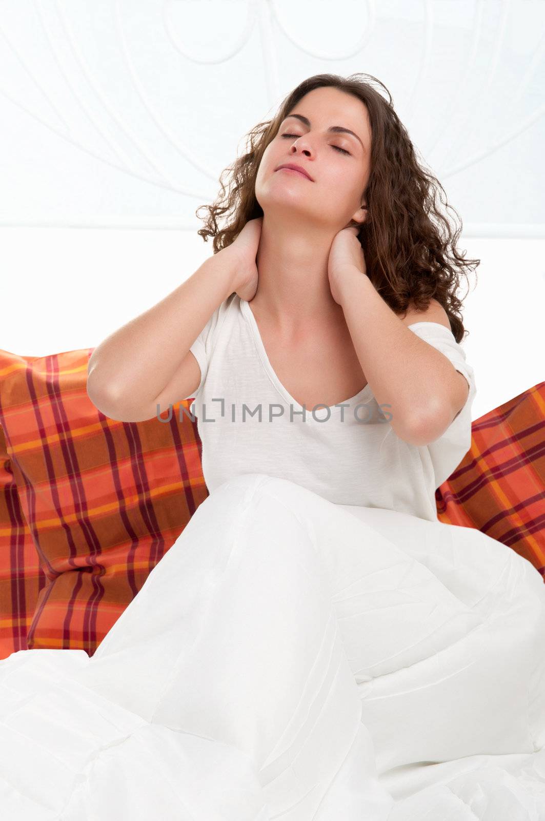 Young woman sitting in her bed after waking up, stretching and holding her neck