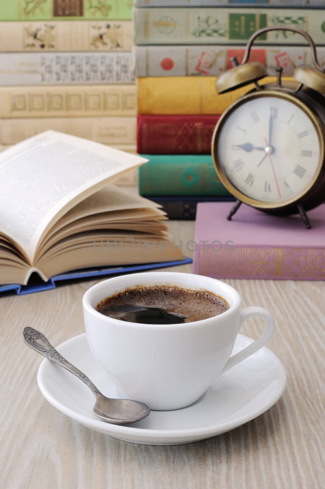 A cup of coffee on the table against the background of an open book with a clock and a stack of books