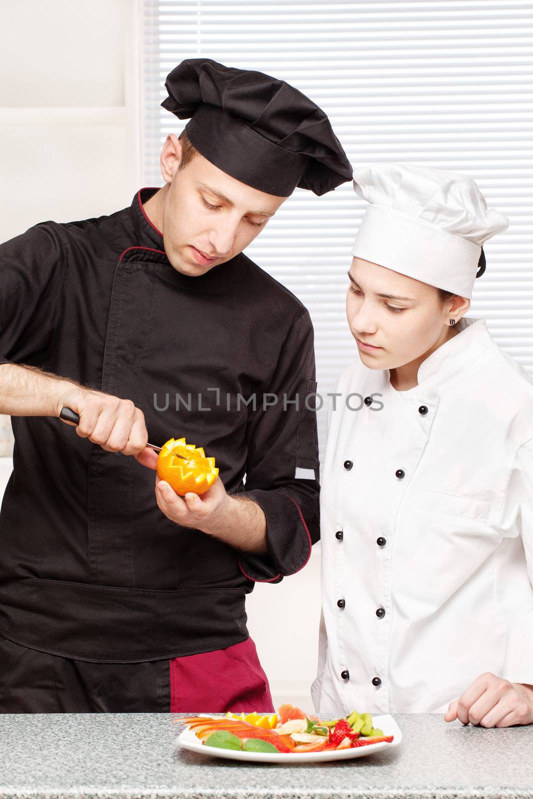 Senior chef teaches young chef to decorate fruit plate in kitchen