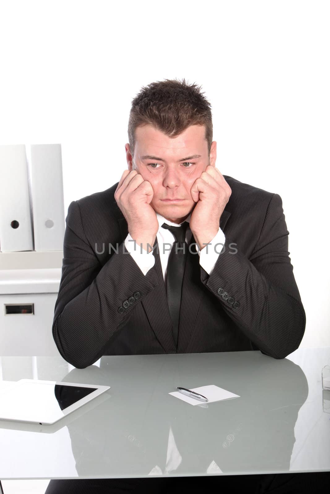 Depressed businessman sitting at his desk with his head resting on his hands staring glumly into the distance