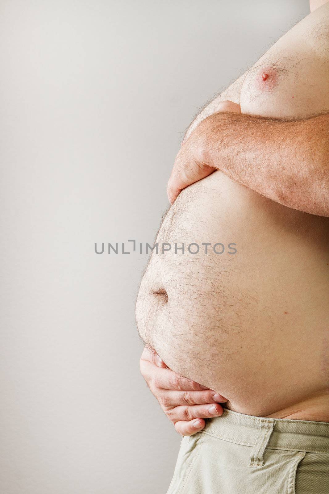 A Man holding his belly like a pregnant Woman. Man is topless with hands supporting both the top and bottom of his beer belly.