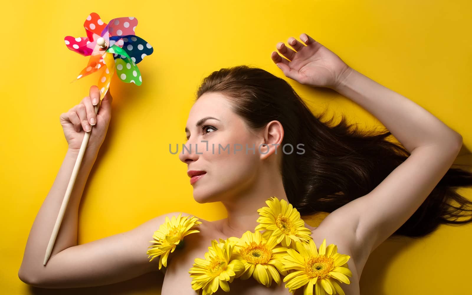 Topless portrait of a beautiful young woman covered with yellow flowers holding colorful pin wheel isolated on yellow background