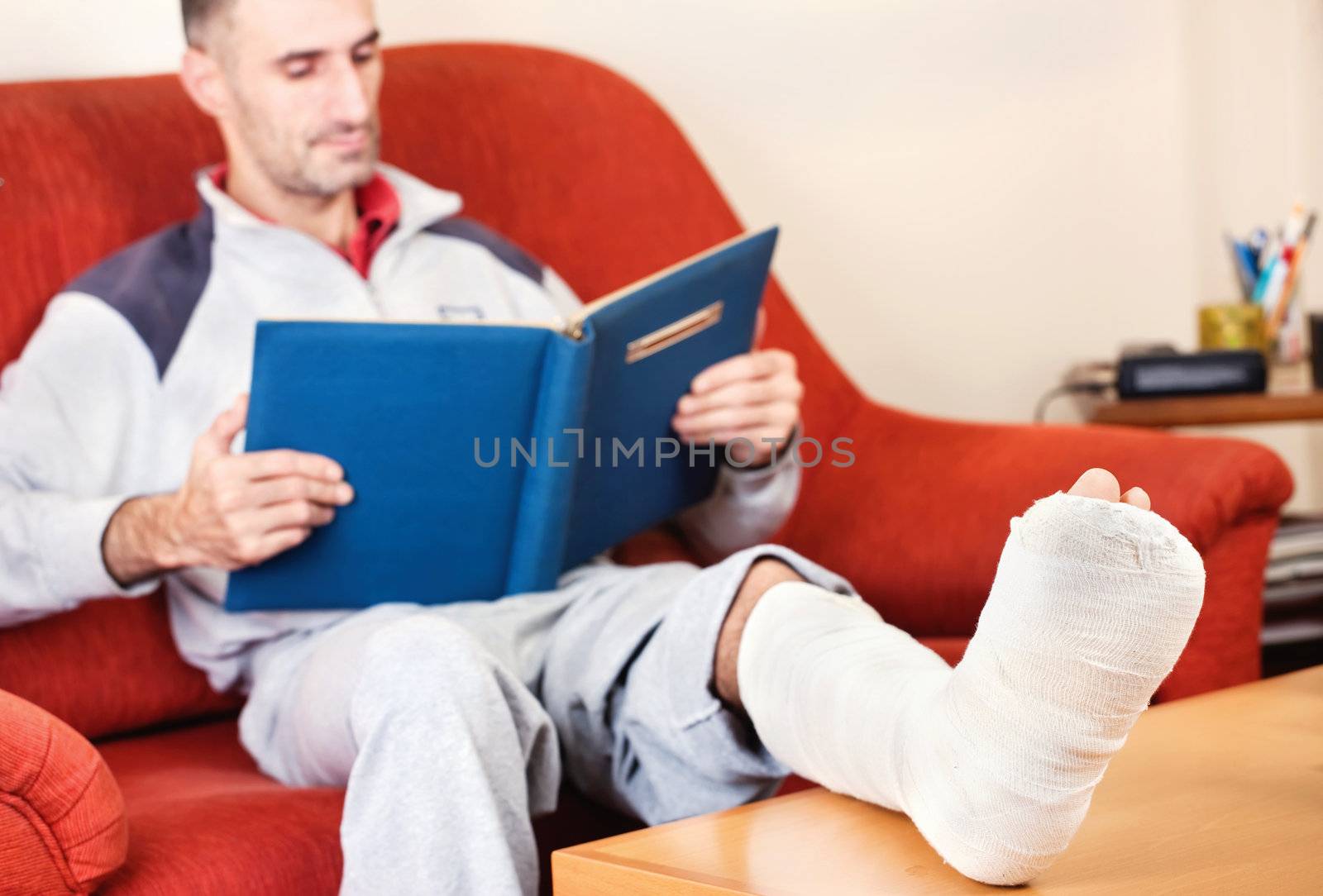 man with a broken leg on a sofa at home  reading book; focus on cast