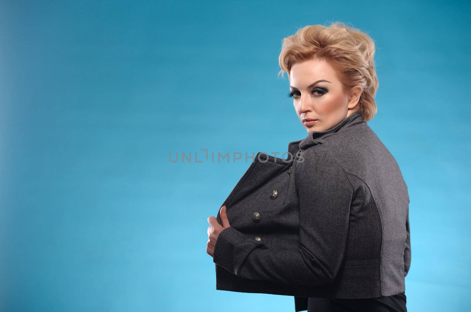 Portrait of beautiful blond femme fatale with shirt hair in gray jacket and black dress posing on blue background