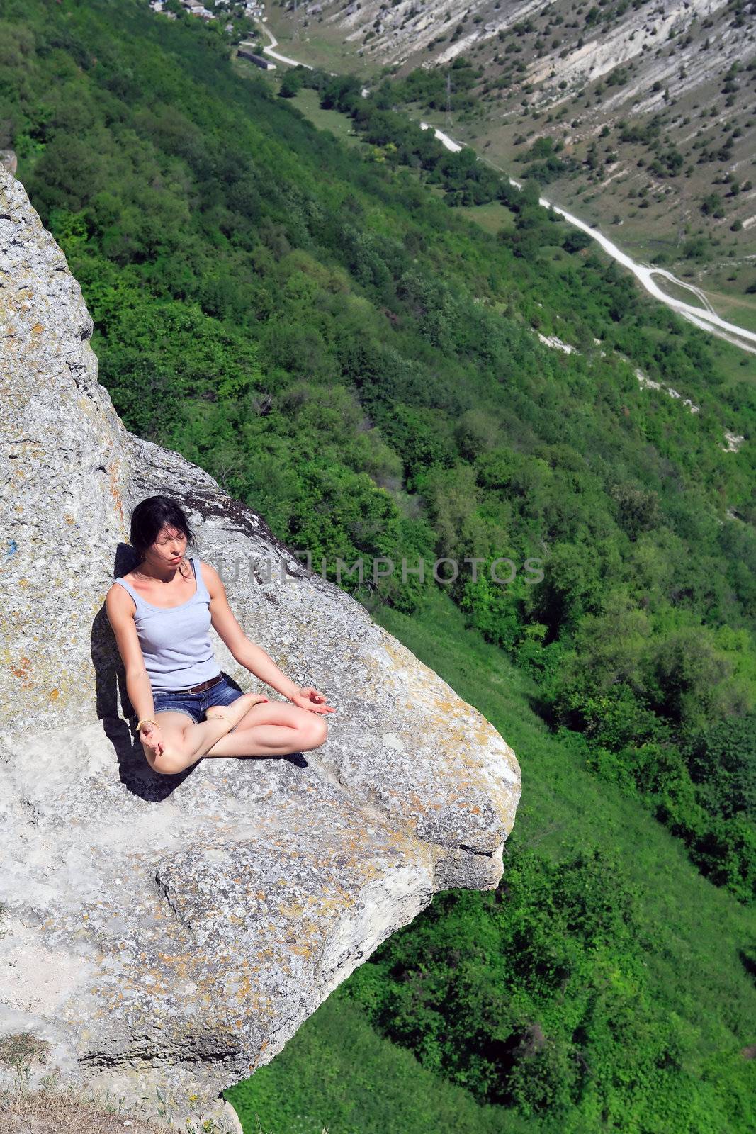 Beautiful adult woman sitting in lotus position on the rock above precipice