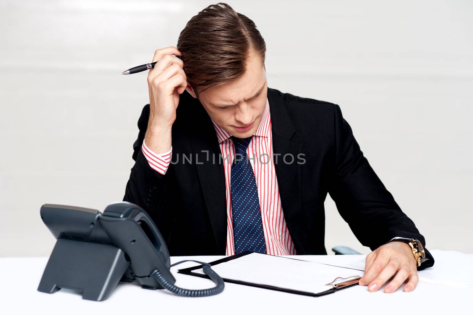 Confused young man itching his head with pen. Sitting at work desk