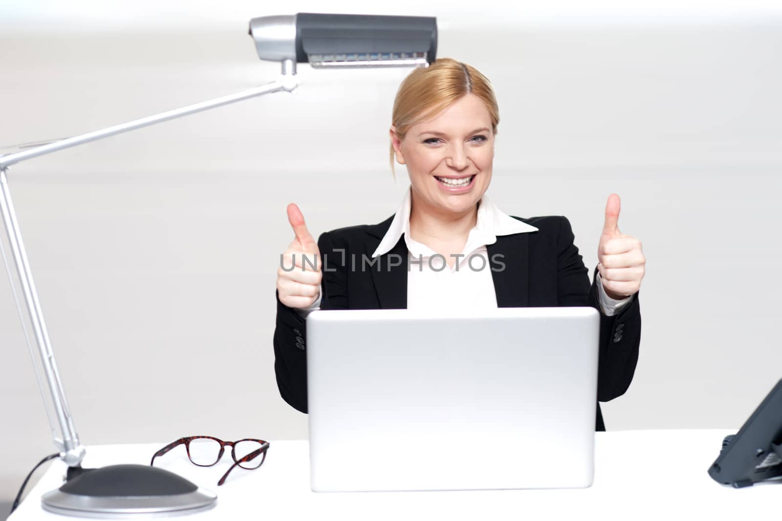 Cheerful businesswoman in office with laptop gesturing double thumbs up