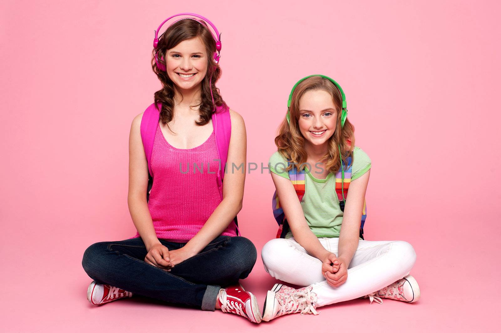 Two school friends sitting on studio floor. Carrying school bag and listening to music