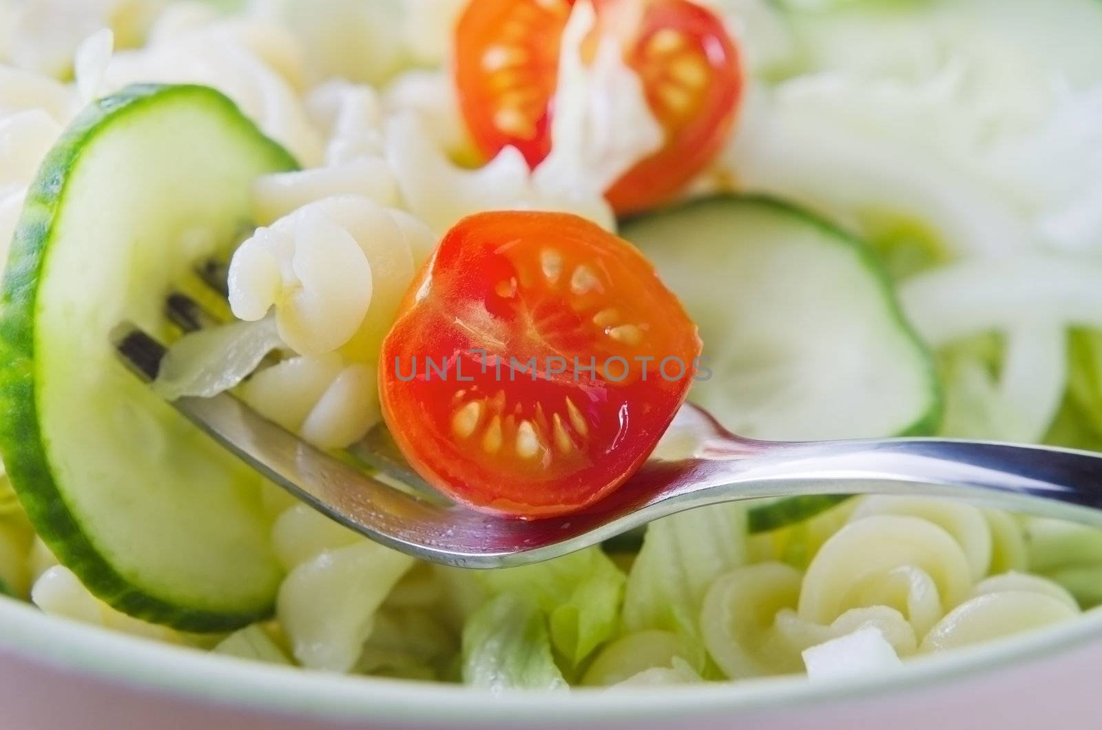 Close up (macro) of a fork, lifting a red cherry tomato, pasta and cucumber slice from a bowl of green salad, with tomato in centre of frame.