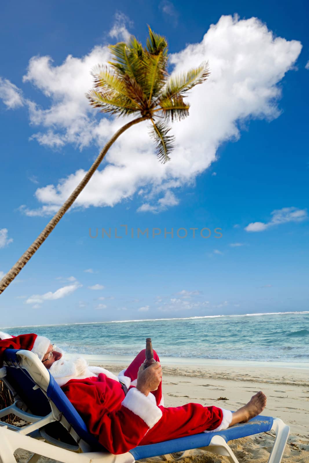 Santa claus is on vacation. He is resting on a sun lounger on exotic beach drinking a beer.