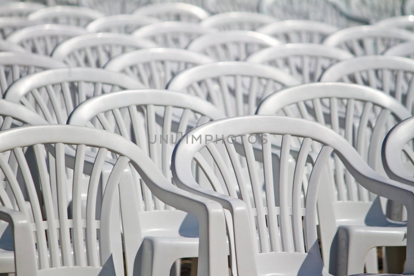 View of some white plastic cafe, restaurant or garden chairs, in an aligned fashion on a outdoor setting.