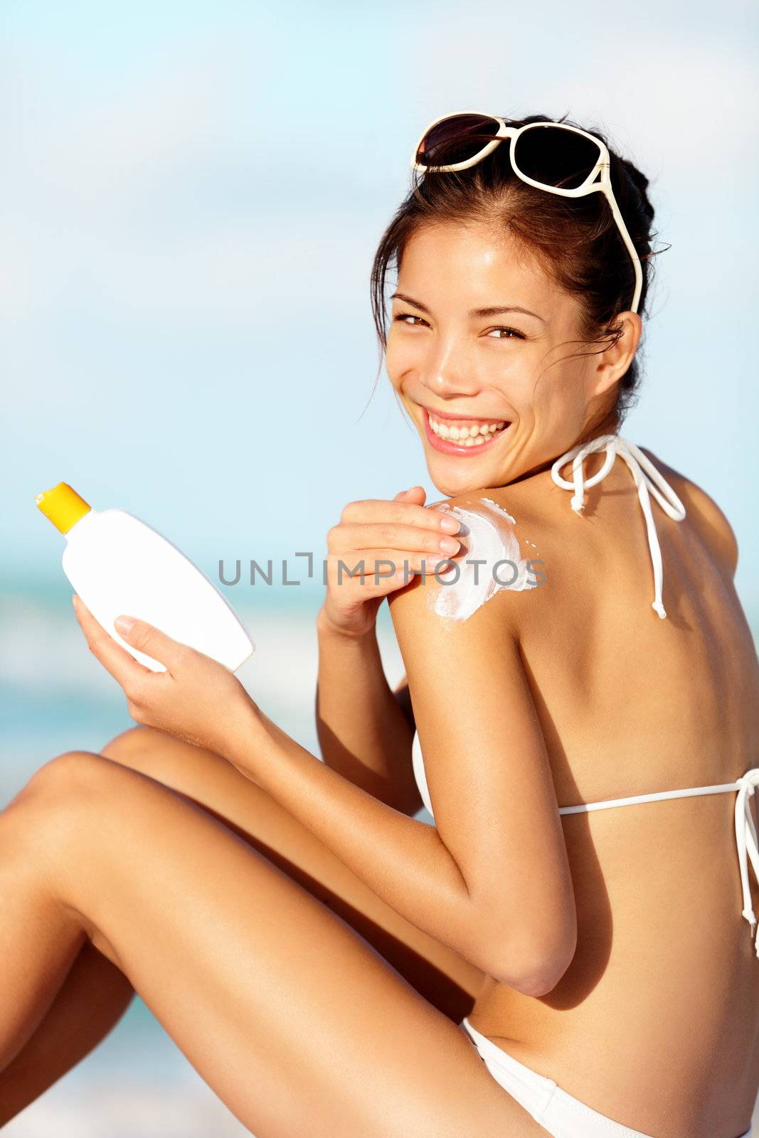 Sunscreen woman. Girl putting sun block lotion on sunny beach holding white sun tan lotion bottle. Beautiful young woman enjoying sunshine on sunny summer day during vacation. Mixed race Asian Chinese / Caucasian female bikini model.