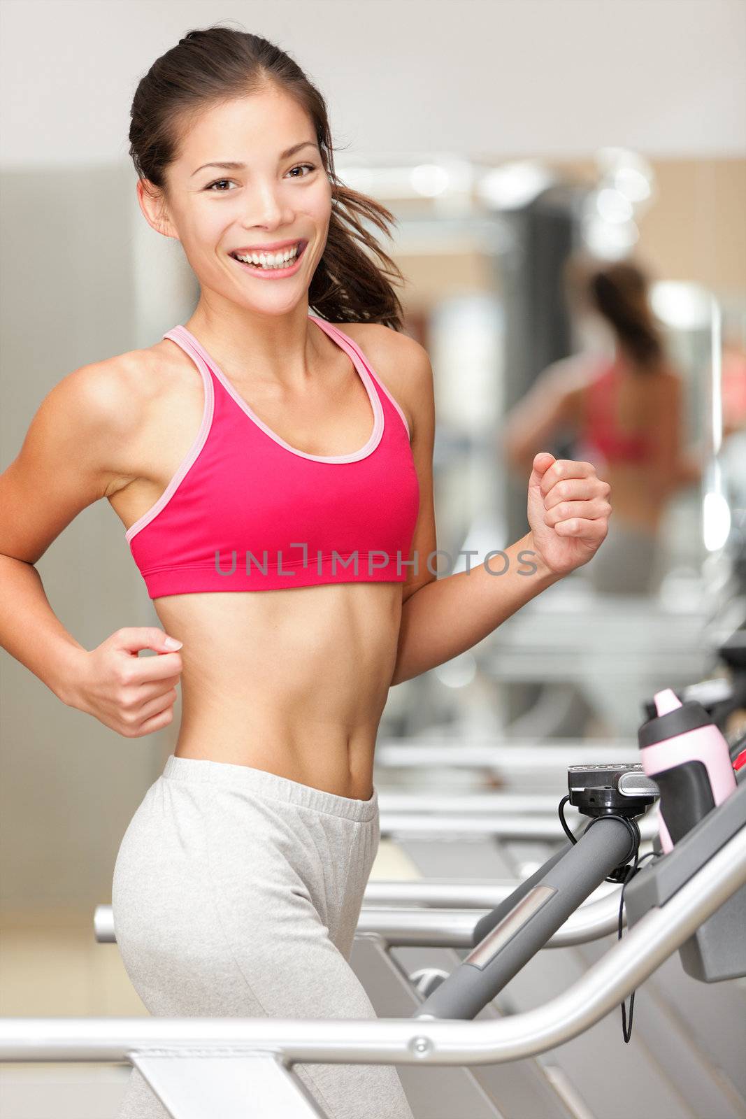 Woman running on treadmill in gym. Fitness model jogging indoors in health club smiling happy. Beautiful young mixed race female model of Chinese Asian and Caucasian ethnicity.