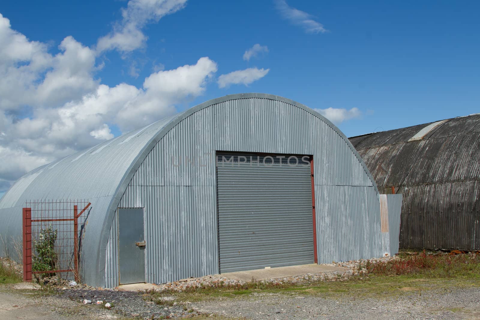 An industrial unit made from corrugated metal sheets with a roller shutter and door on rough ground.