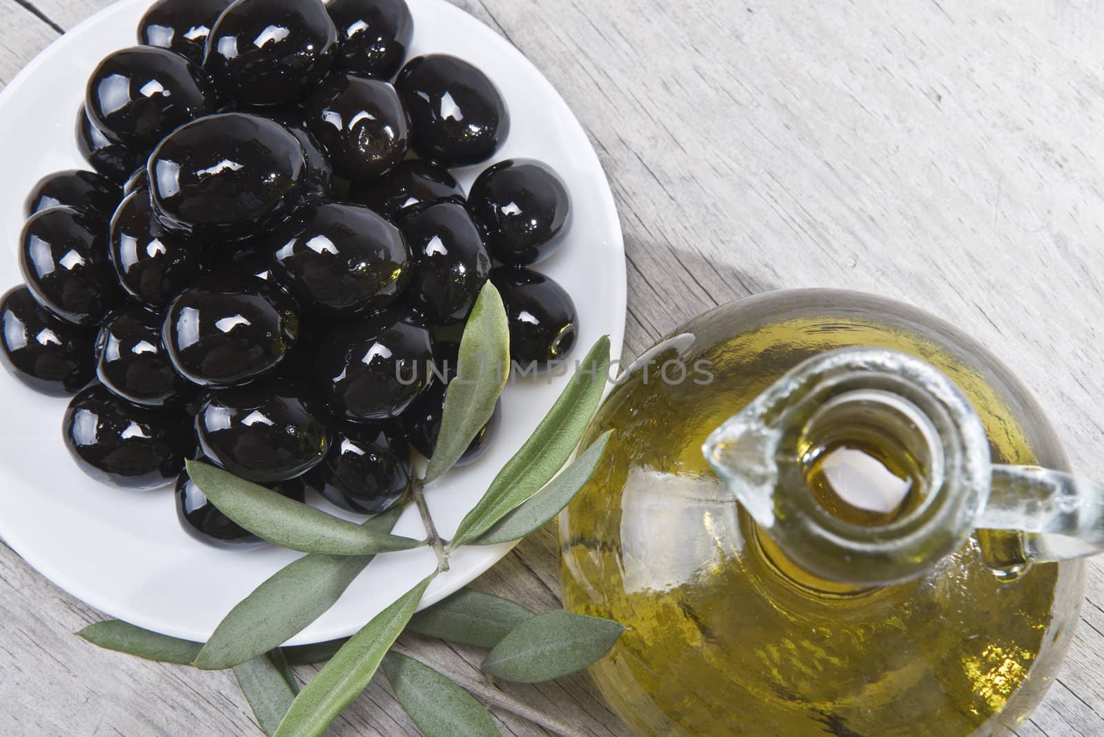 A jar with olive oil ans a plate with black olives on a wooden surface.