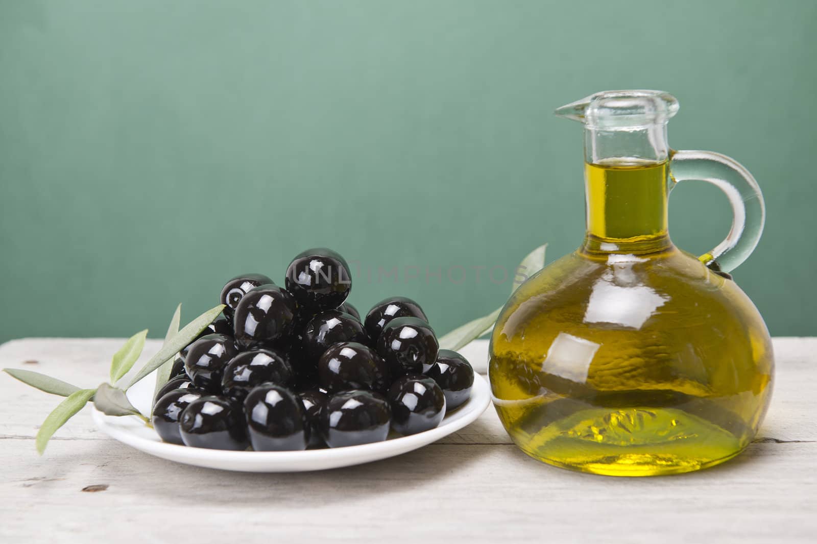 A jar with olive oil ans a plate with black olives on a wooden surface.