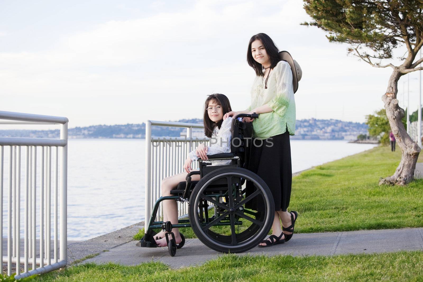 Young teen girl pushing sister in wheelchair outdoors by lake