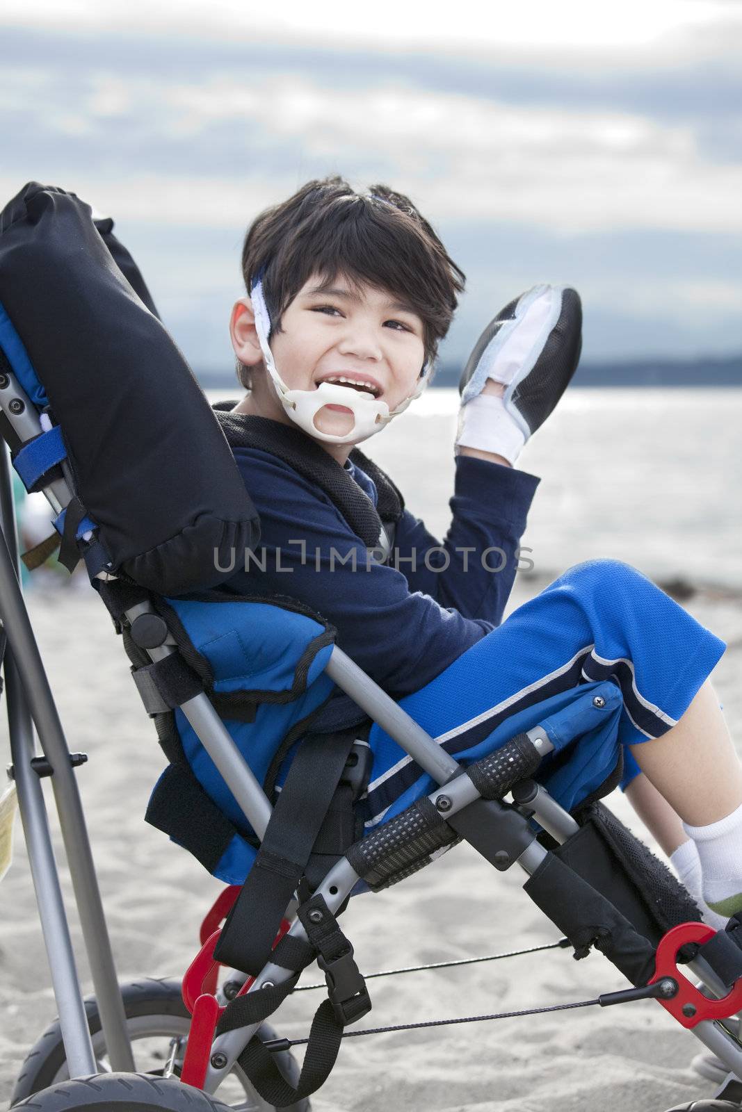 Happy disabled five year old boy in wheelchair on the beach, waving hello