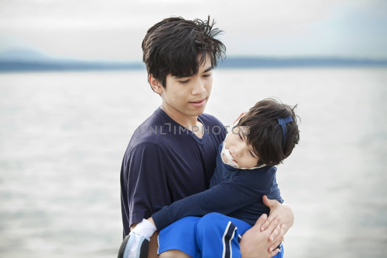 Big brother carrying disabled boy on beach by water. Child has cerebral palsy