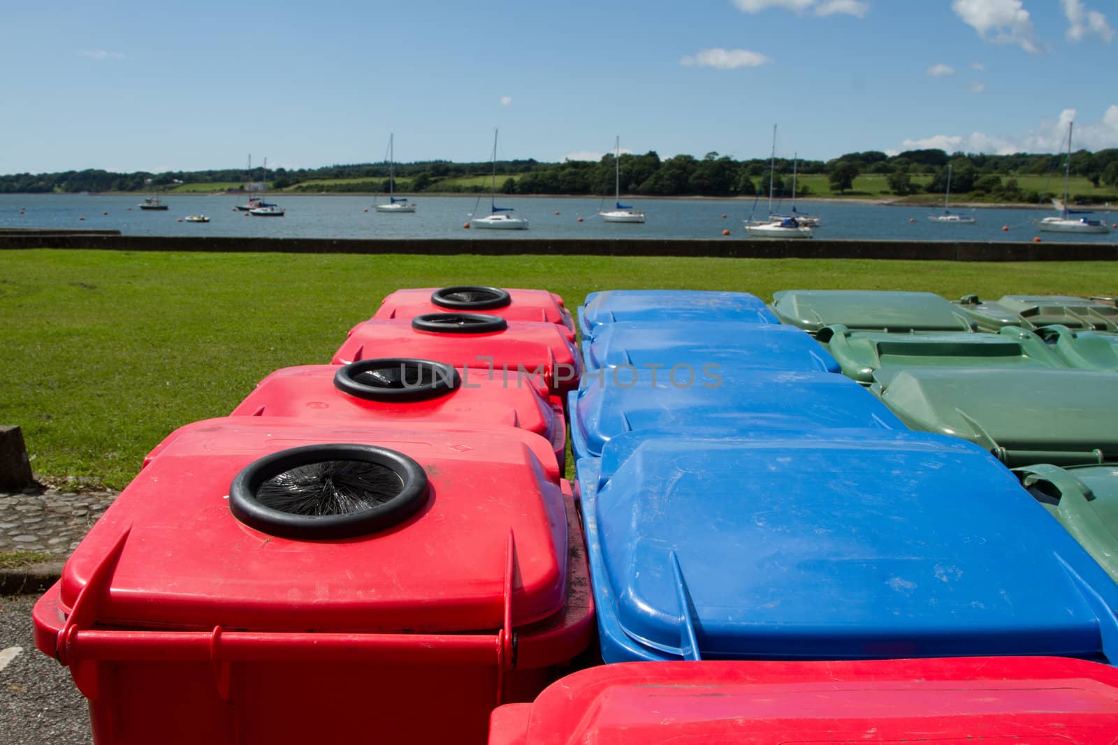 A group of plastic multi-coloured wheelie bins arranged on tarmac infront of a park and a stretch of water with yachts.