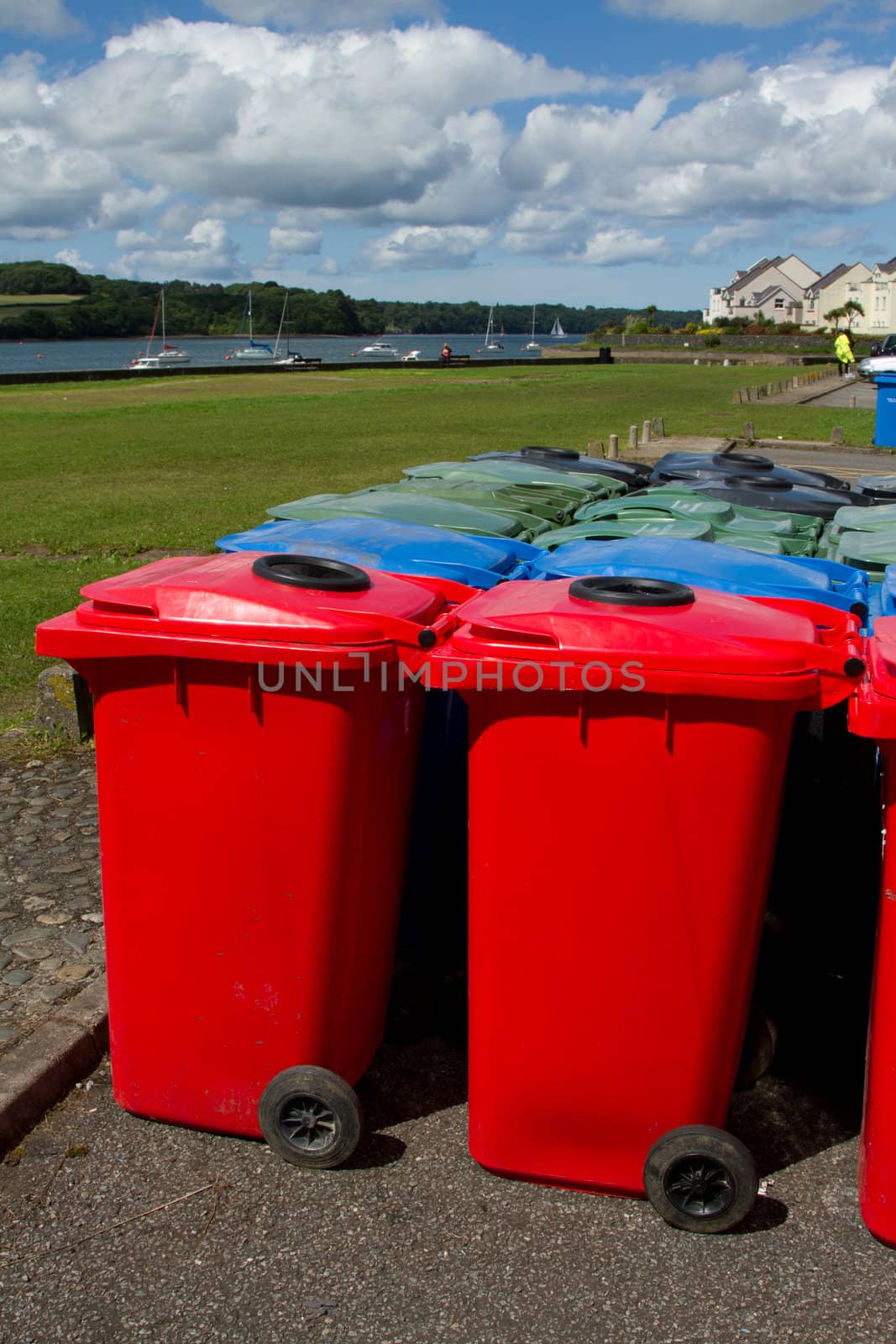 A group of plastic multi-coloured wheelie bins arranged on tarmac infront of a park and a stretch of water with yachts.