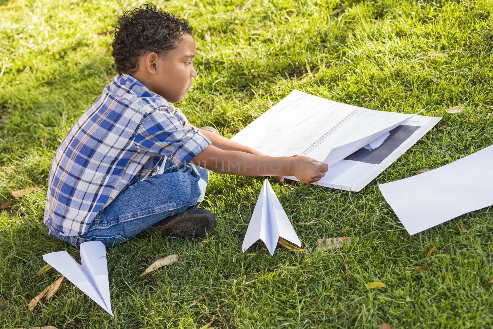 Mixed Race Boy Learning How to Fold Paper Airplanes Outdoors on the Grass.