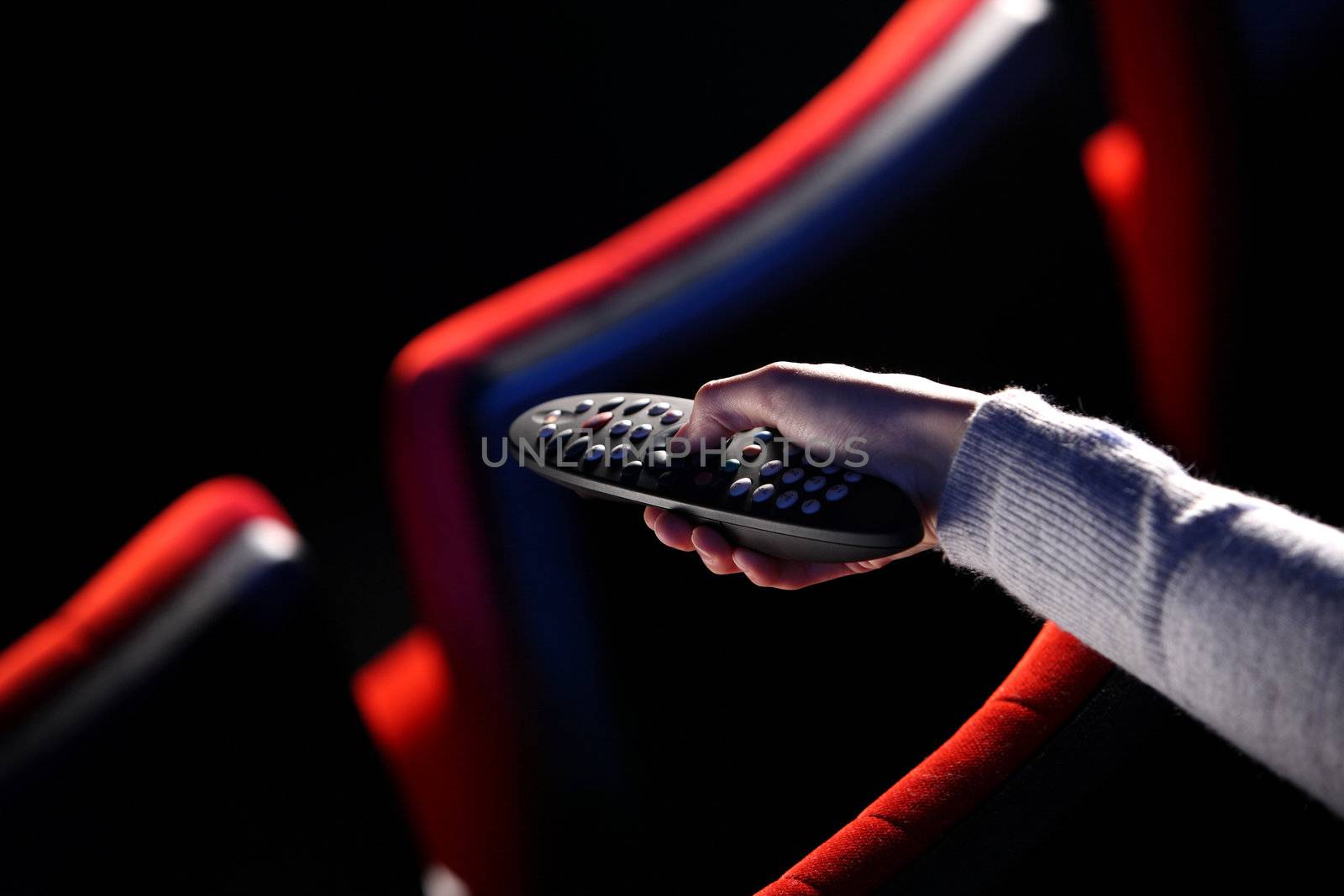 closeup of a hand holding a remote control TV, in the background you can see the red chairs in a movie theater. conceptual image