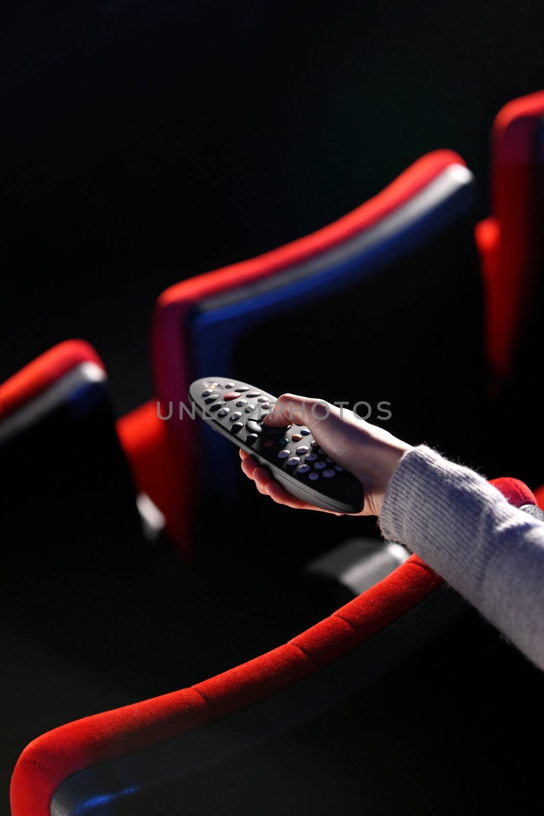 closeup of a hand holding a remote control TV, in the background you can see the red chairs in a movie theater. conceptual image