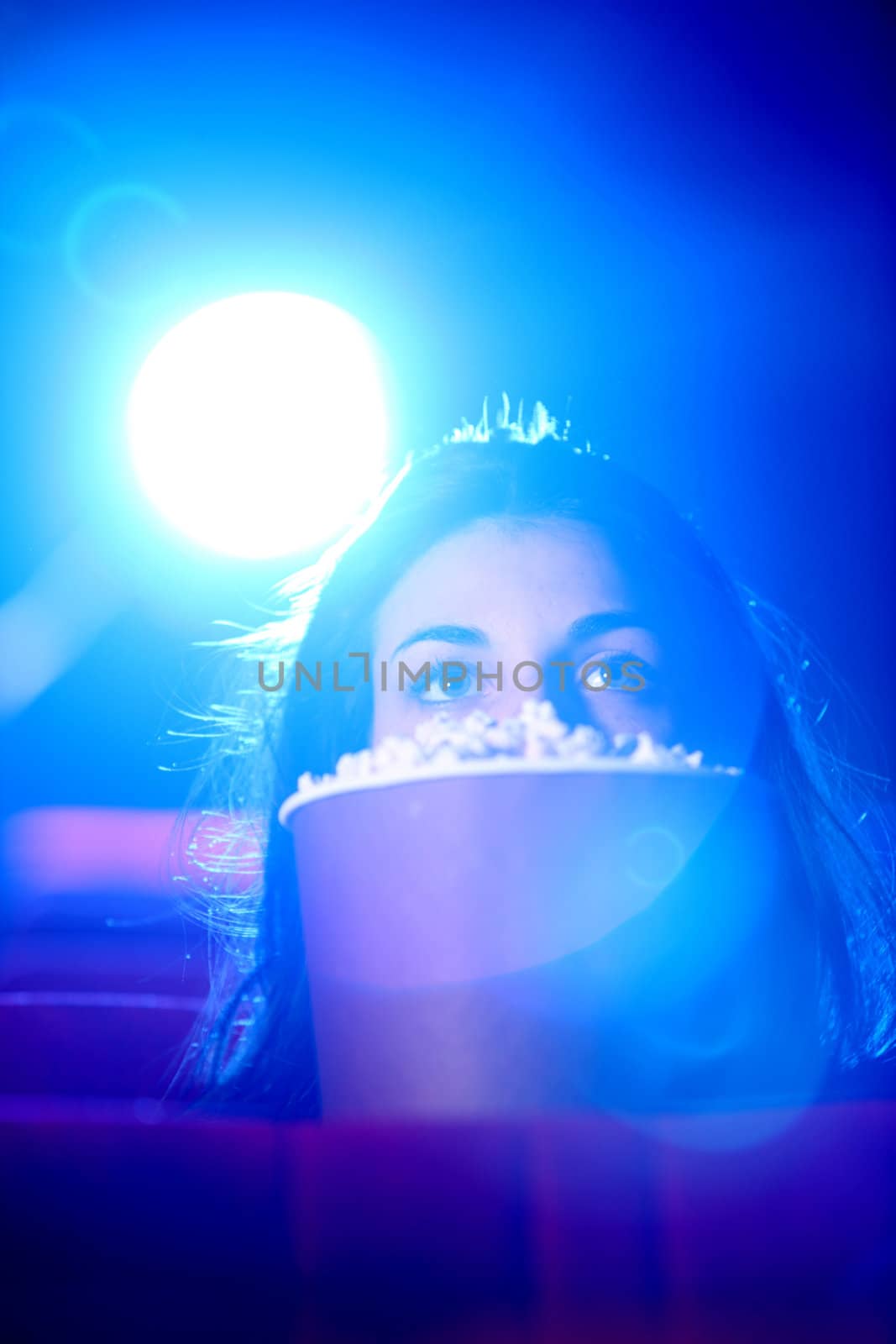 
a pretty young woman sitting at theater, she is scared, and hides behind a box of popcorn