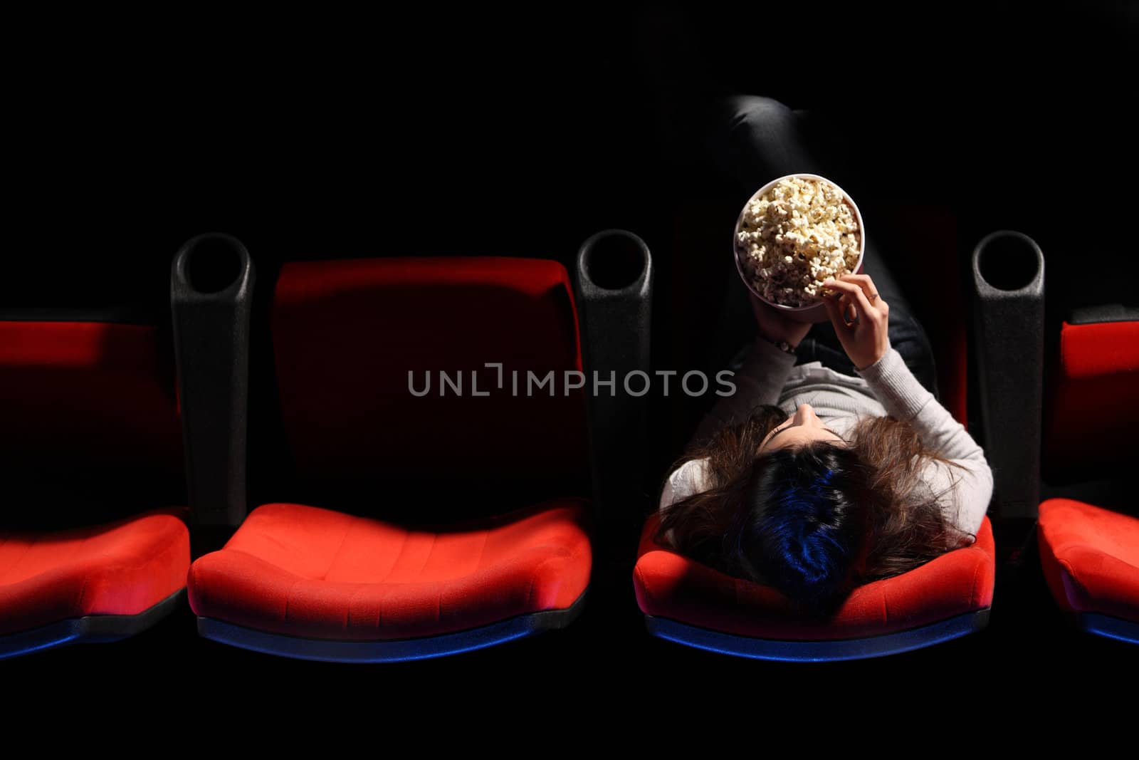 a pretty young woman sitting in a row of red chairs in a theater, she eats popcorn, top view
