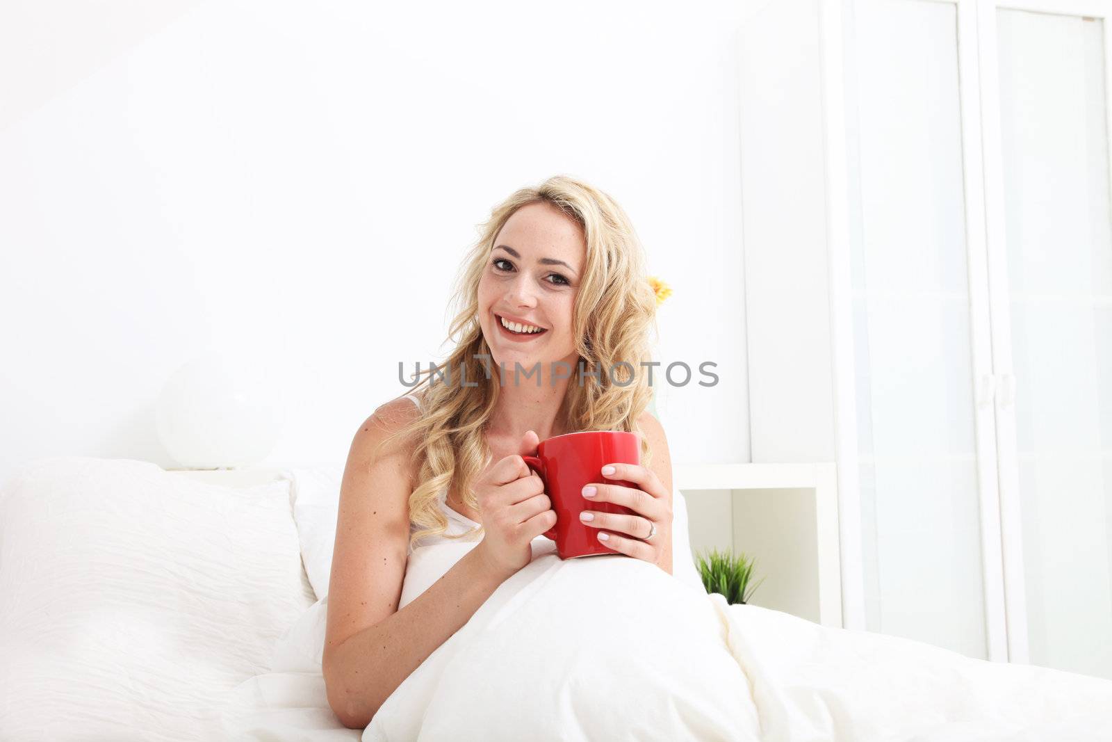Friendly woman with beautiful smile cuddled up in her nice warm bed enjoying an early morning cup of coffee