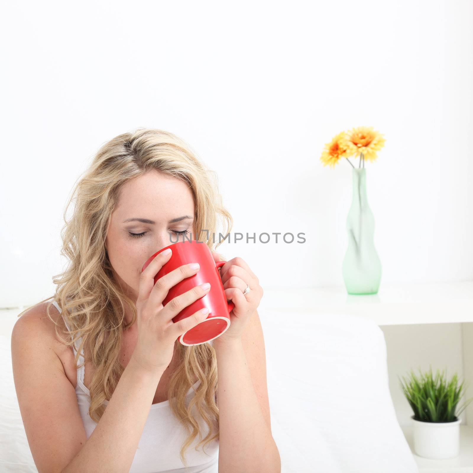 Young woman with long blonde curly hair drinking a large red mug of coffee in bed