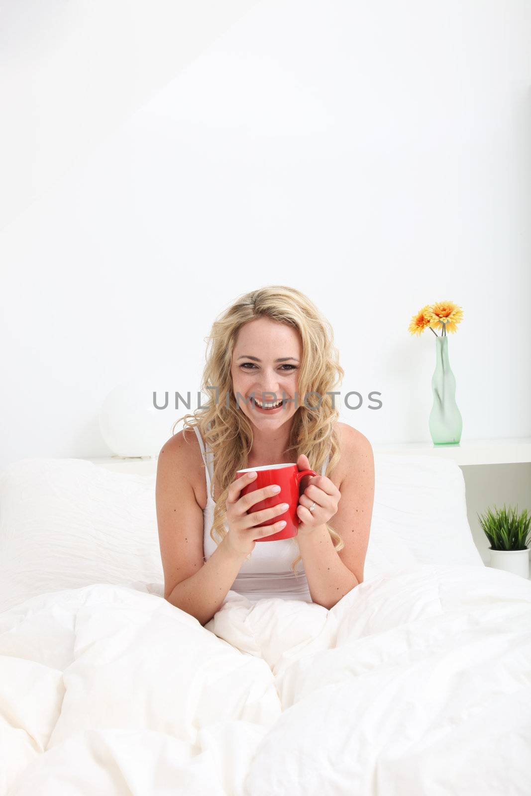 Vivacious young woman with lovely curly blonde hair enjoying a cup of coffee while sitting in bed