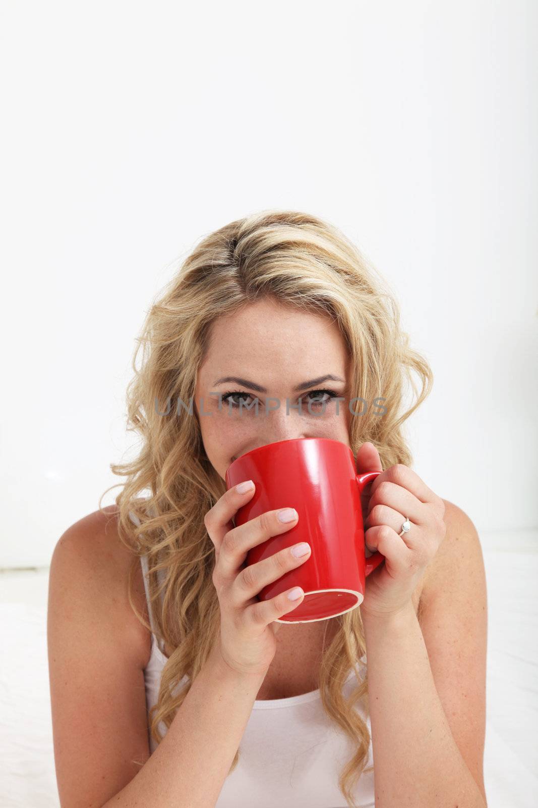 Young blonde woman with smiling eyes drinking a large mug of coffee against a white interior background