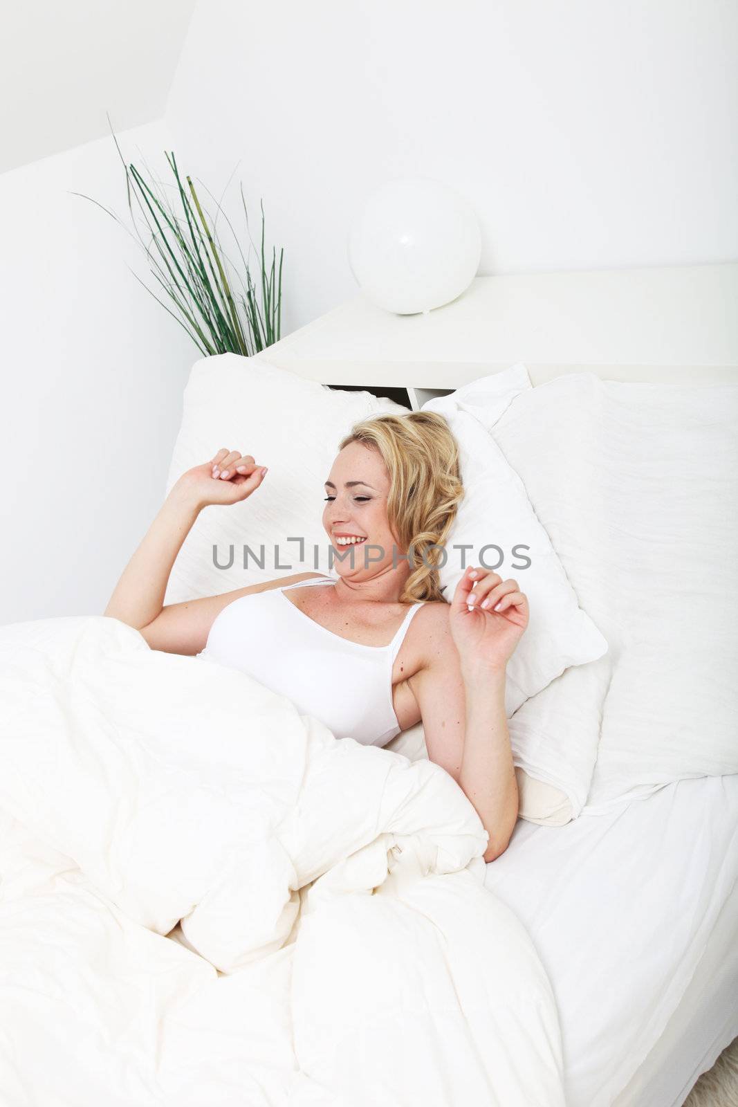 Pretty blonde woman smiling in satisfaction as she stretches to start the day while reclining on her pillows in bed