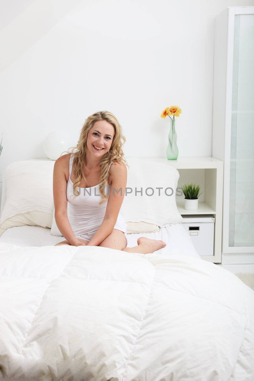 Friendly blonde woman sitting on her bed in a white bedroom interior