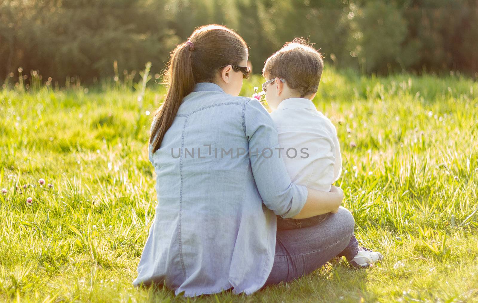 Back view of cute son giving a bouquet of flowers to his mother sitting in a sunny field