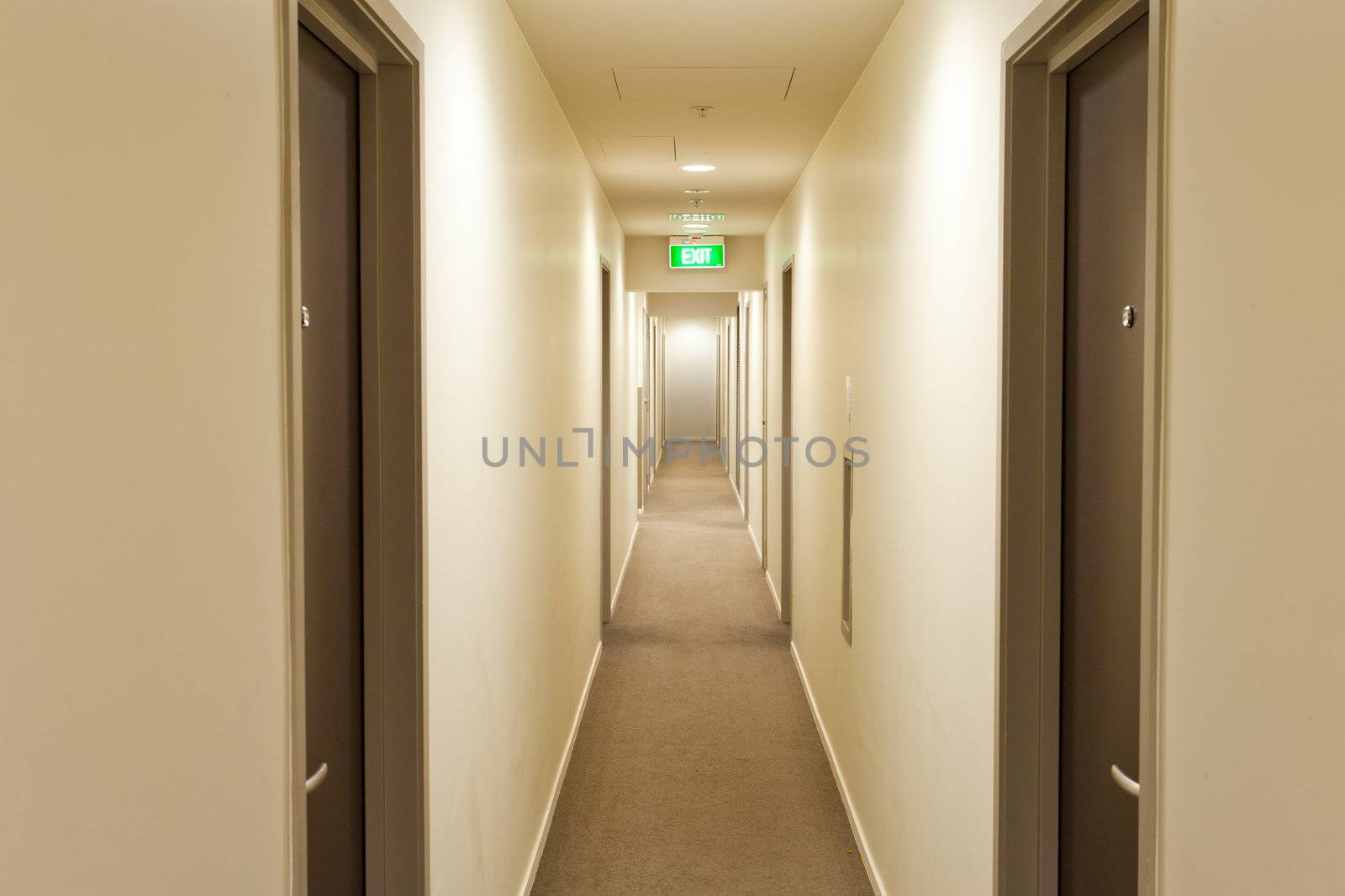 Long empty corridor in a hotel or apartment building interior with overhead lighting, doors on either side and illuminated exit sign