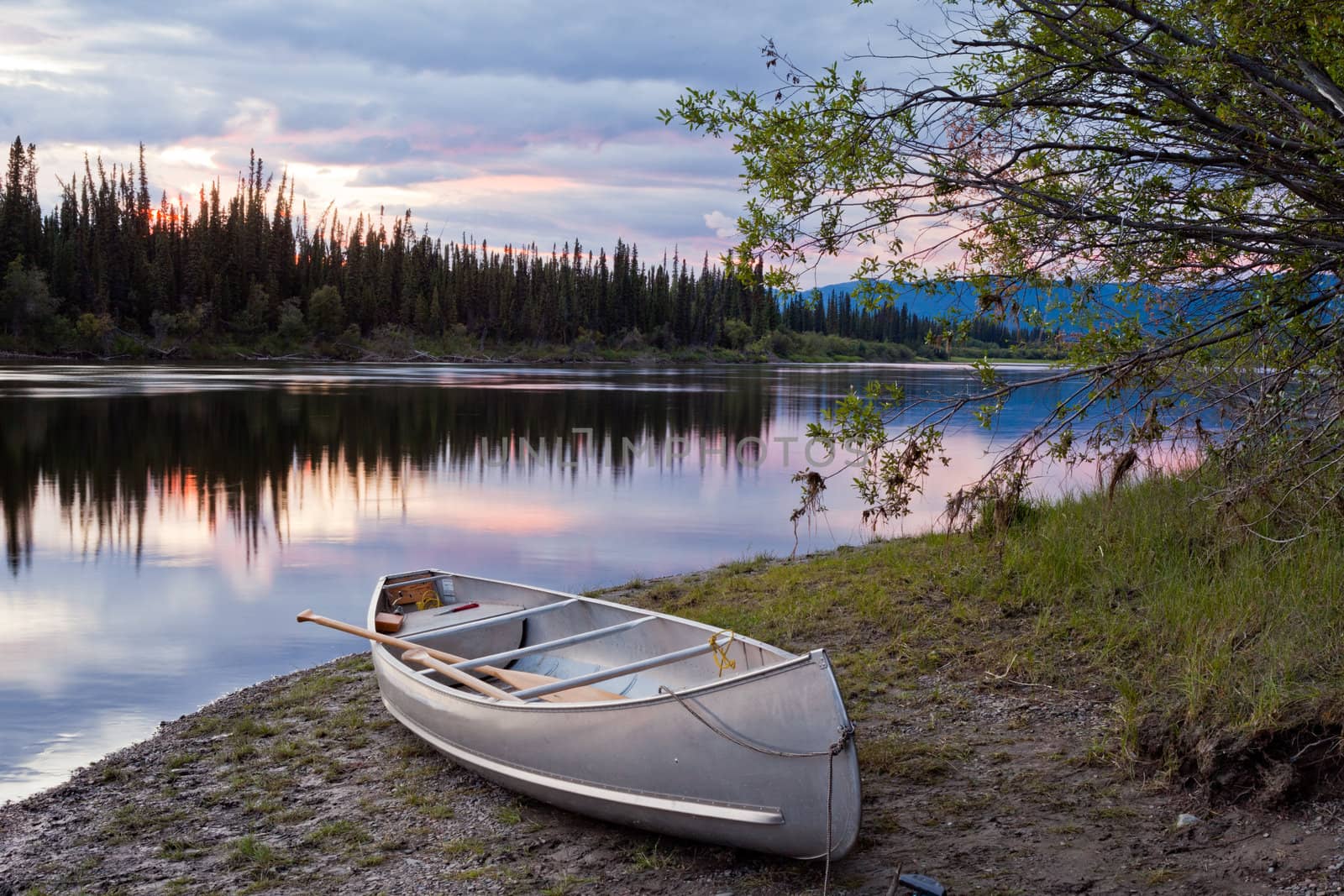 Canoe and paddles beached on shore of beautiful Teslin River in the remote wilderness of Yukon Territory, Canada, the river surface reflecting delicate sunset colors