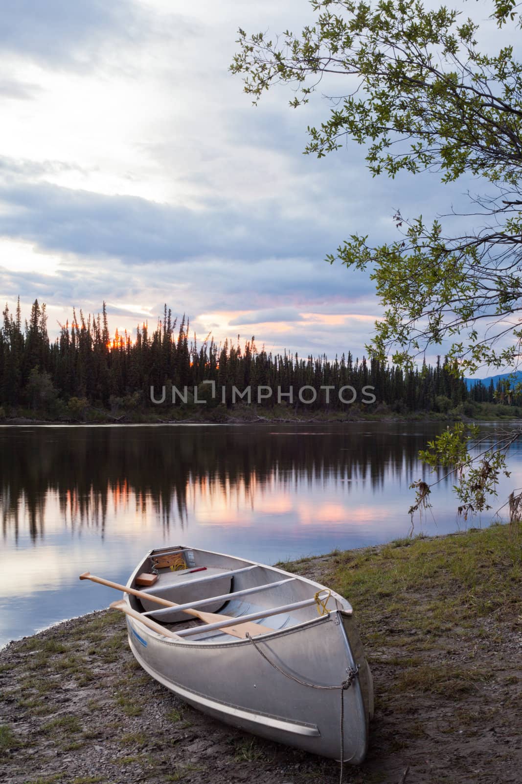 Canoe and paddles beached on shore of beautiful Teslin River in the remote wilderness of Yukon Territory, Canada, the river surface reflecting delicate sunset colors