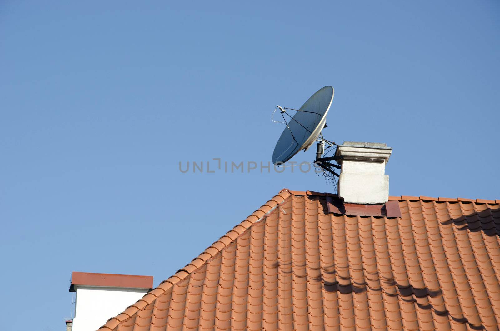Satellite television antenna attached to chimney on red tiled roof house on background of blue sky.